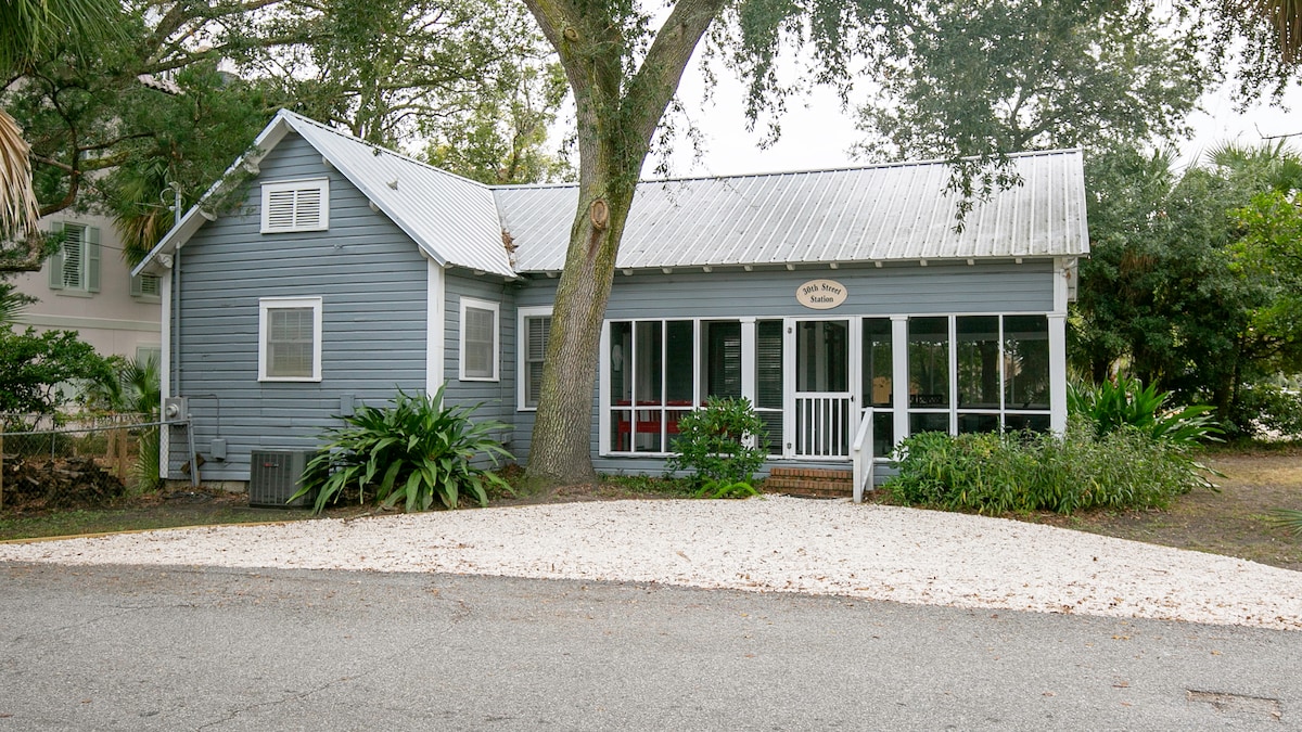 A historic cottage with light blue siding and a metal roof is surrounded by lush greenery. The welcoming front porch features a white railing and a screened area. A spacious gravel driveway leads up to the entrance, providing ample space for parking.