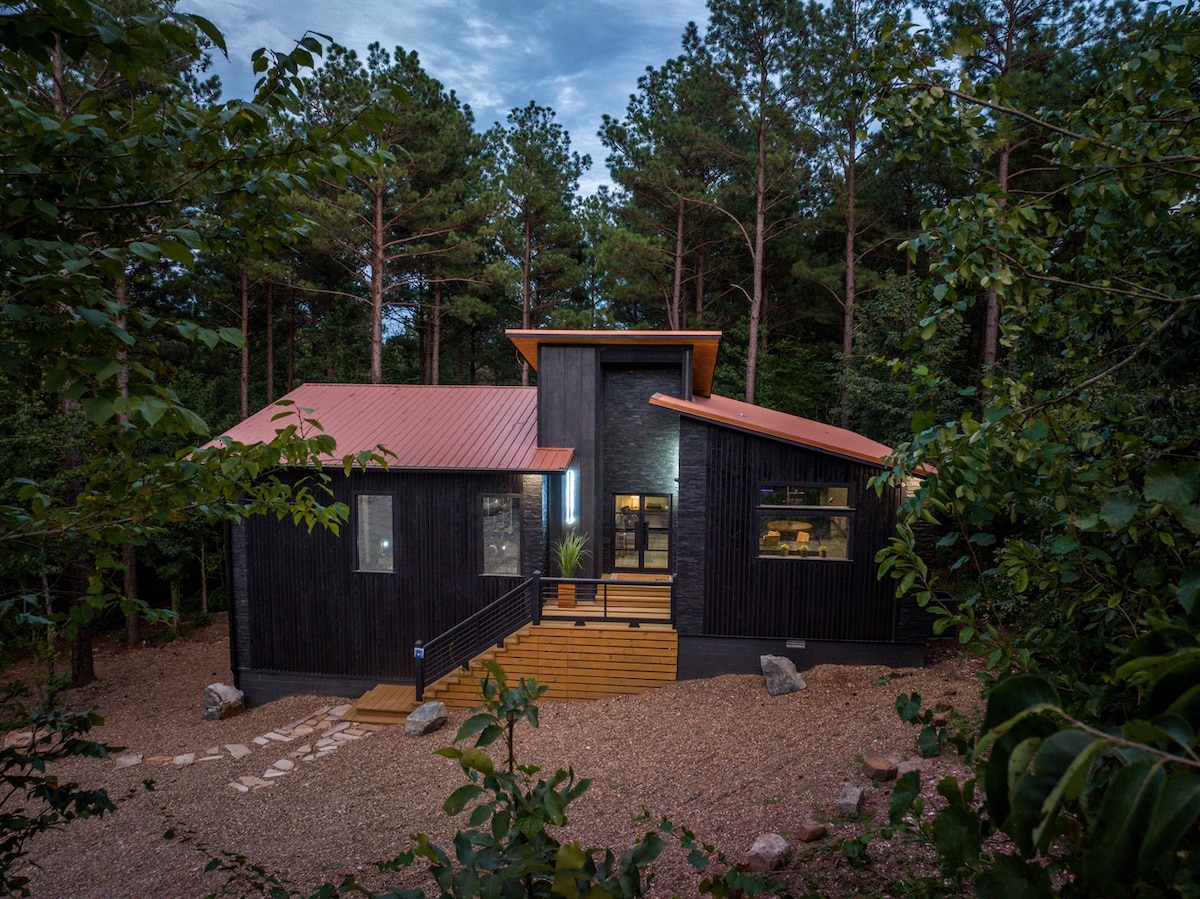 A contemporary cabin is set amidst lush greenery, showcasing a striking black exterior with a striking red roof. Stone pathways lead to the entrance, framed by steps that enhance the inviting facade. Tall trees and natural landscape surround the property, creating a serene atmosphere.