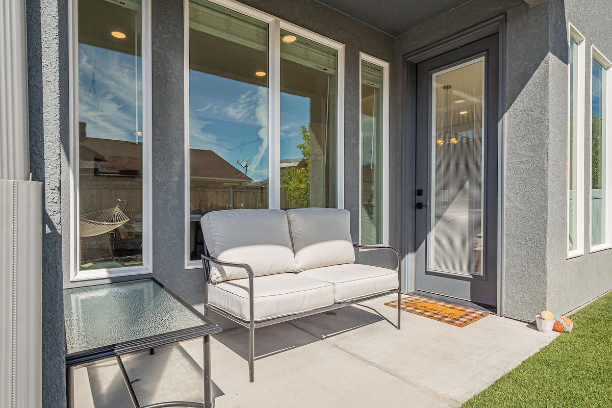 A cozy outdoor sitting area is presented, featuring a cushioned loveseat and a glass-top table. Large windows surround the space, allowing natural light to brighten the area. A welcome mat lies in front of the door, complementing the inviting atmosphere.