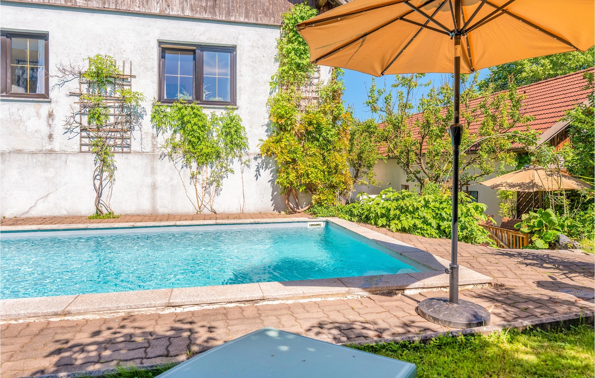 An outdoor pool is surrounded by lush greenery, creating a serene space for relaxation. A large orange umbrella provides shade next to the poolside, while a small table is situated nearby. The building's white wall is complemented by climbing plants and multiple windows.
