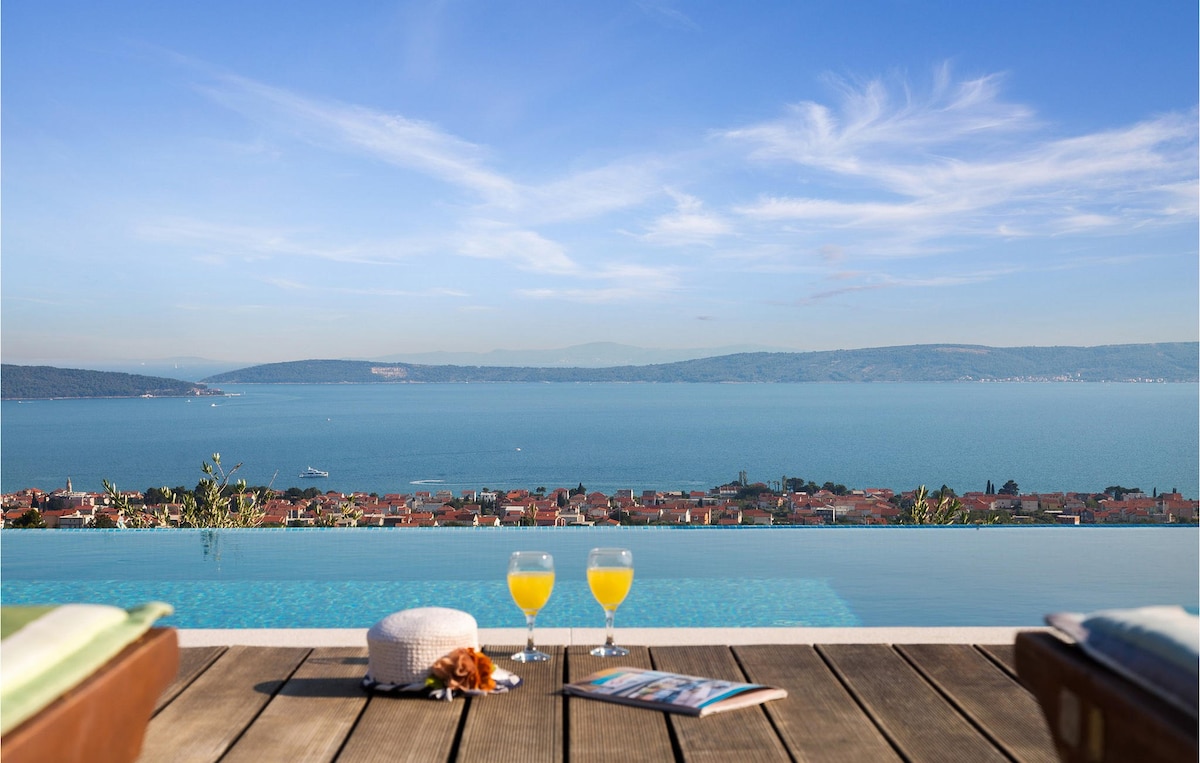 A stunning view over the sea and coastline is presented from the edge of a large infinity pool. Two glasses of orange juice are positioned on a wooden deck, accompanied by a hat and a magazine, creating a serene atmosphere for relaxation.