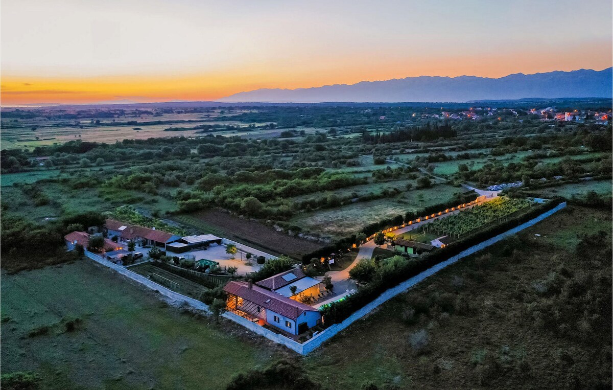 An aerial view captures the expansive villa and its surroundings at sunset, highlighting the illuminated pathways and rustic stone walls. The lush landscape stretches into the distance, bordered by mountains under a soft twilight sky, offering a sense of tranquility and privacy.