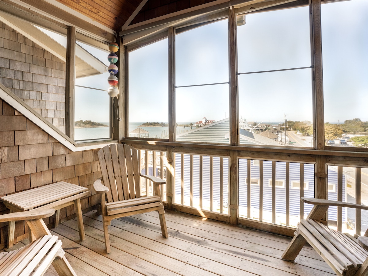 A furnished screened porch features wooden Adirondack chairs and a small table, providing comfortable seating. Large windows allow for natural light and views of the surrounding area, enhancing the space's connection to the outdoors.