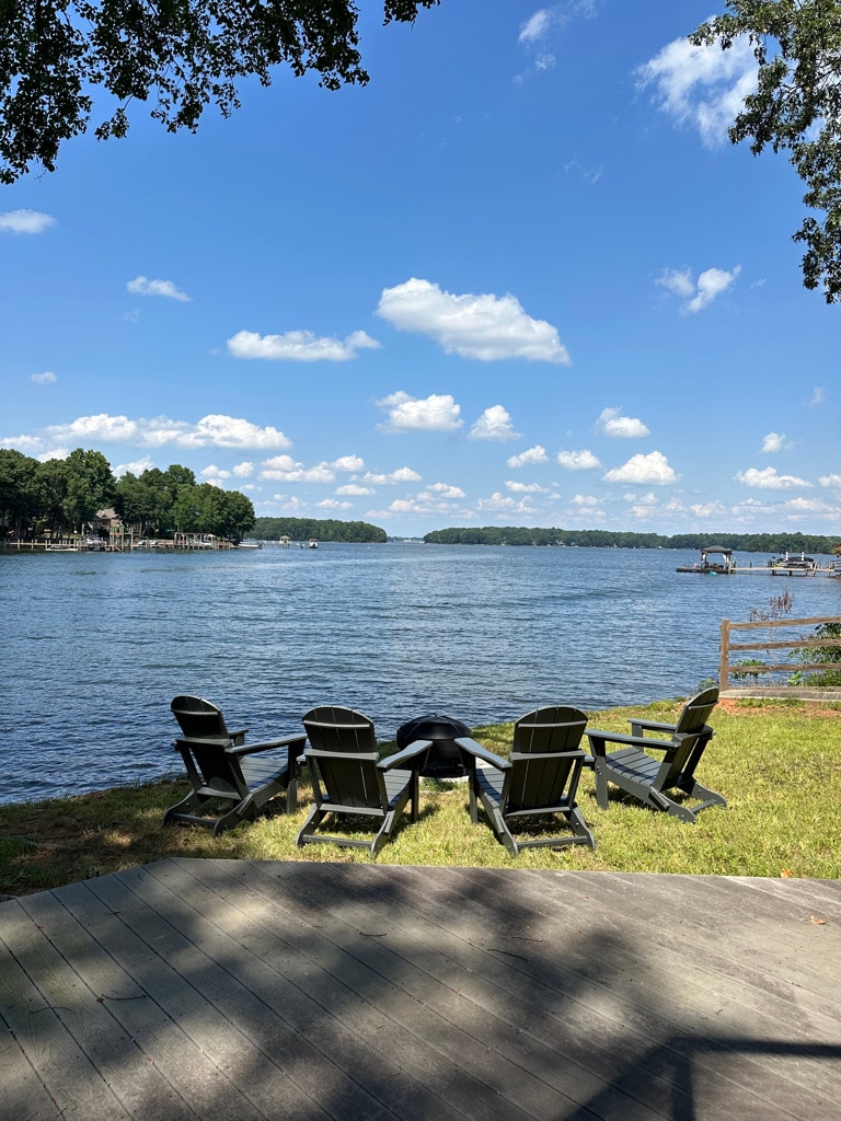 Four black Adirondack chairs are arranged facing the tranquil waters of Lake Norman, surrounded by greenery. The sky is bright blue with fluffy white clouds, and the gentle ripples of the lake reflect the sunlight. A wooden deck provides a spacious area for relaxation.