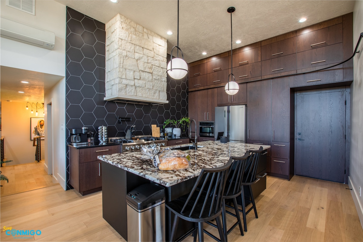 A modern kitchen is highlighted by dark cabinetry and a stylish granite island, which features seating for six. A large hood vent and stone backsplash add texture, while sleek appliances are arranged within easy reach. Natural light illuminates the space through adjacent windows.