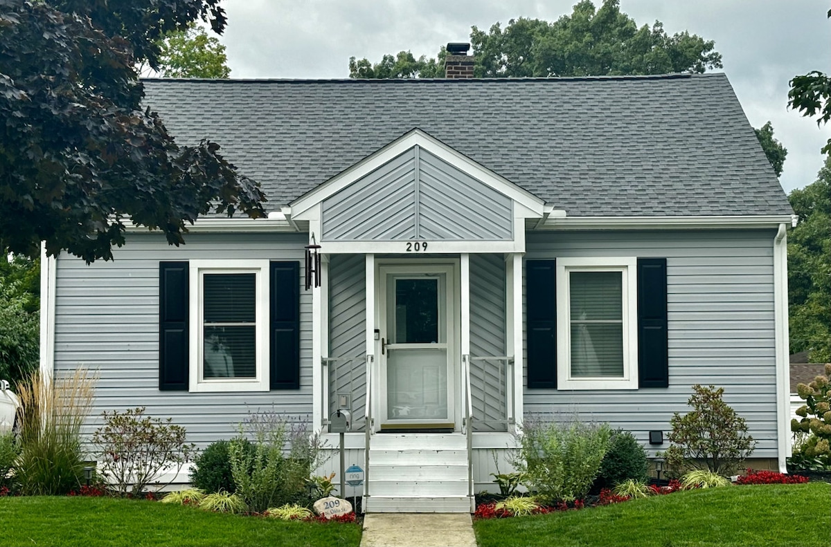A light blue house with white trim is viewed from the front. The entrance features a set of stairs leading to a central door, flanked by two windows. Green landscaping and seasonal flowers add color to the front yard, and a small, subtle porch light brightens the entrance.