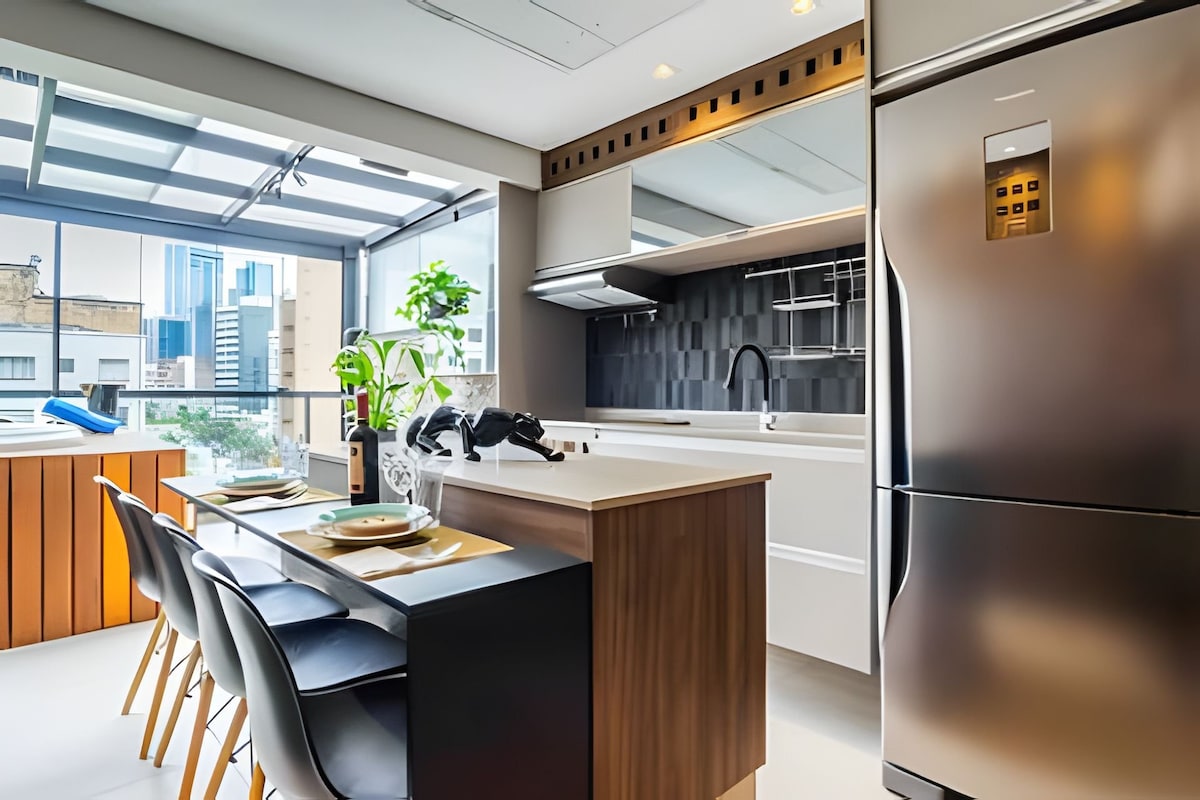 A modern kitchen area includes sleek cabinetry and a stainless steel refrigerator. A breakfast bar with four black chairs is present, accompanied by neutral table settings. Large windows allow for natural light and showcase a view of the cityscape beyond.
