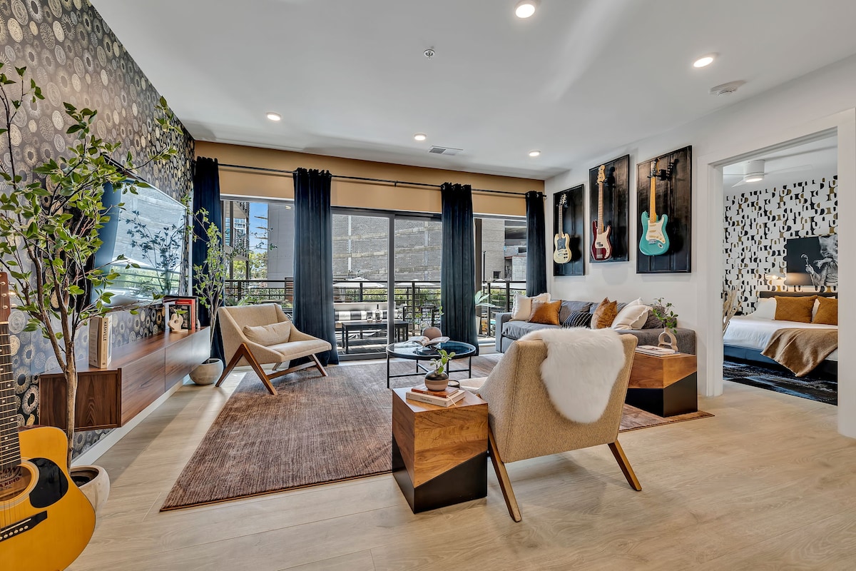 The living area features modern furnishings, including a pair of stylish armchairs and a plush rug. A large window provides natural light and offers a view of the balcony. Custom guitar displays and framed art add character to the walls.