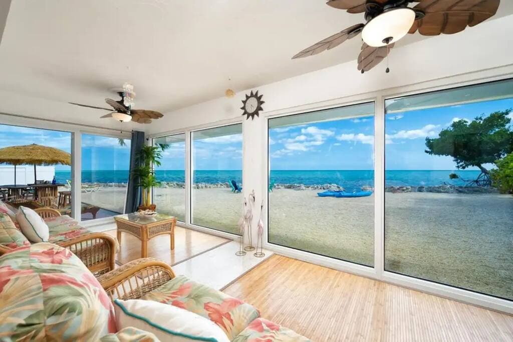 A spacious living area features large glass windows that provide unobstructed views of the ocean, allowing natural light to fill the space. Wicker furniture with colorful cushions invites relaxation, while a decorative coffee table sits at the center, creating a perfect spot for socializing.