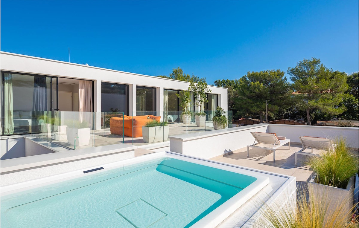 A modern outdoor space features a clear swimming pool beside delicate landscaping. Sun loungers are positioned nearby, providing ample seating. Expansive glass windows and doors extend from the villa, complementing the bright blue sky above and creating a seamless connection between outdoor and indoor areas.