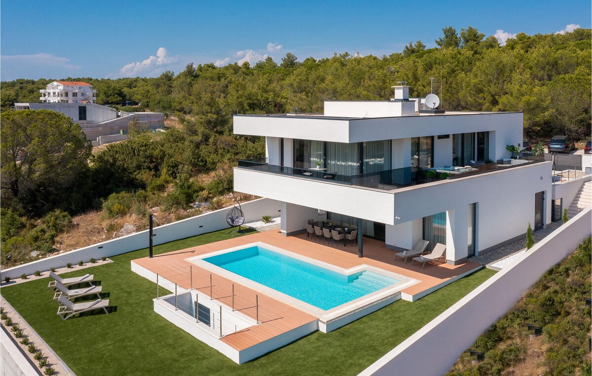 A modern villa is showcased, featuring a spacious outdoor area with a clear swimming pool surrounded by sun loungers. Lush green landscaping contrasts against the villa's sleek white exterior, while large windows invite natural light into the elegant design.