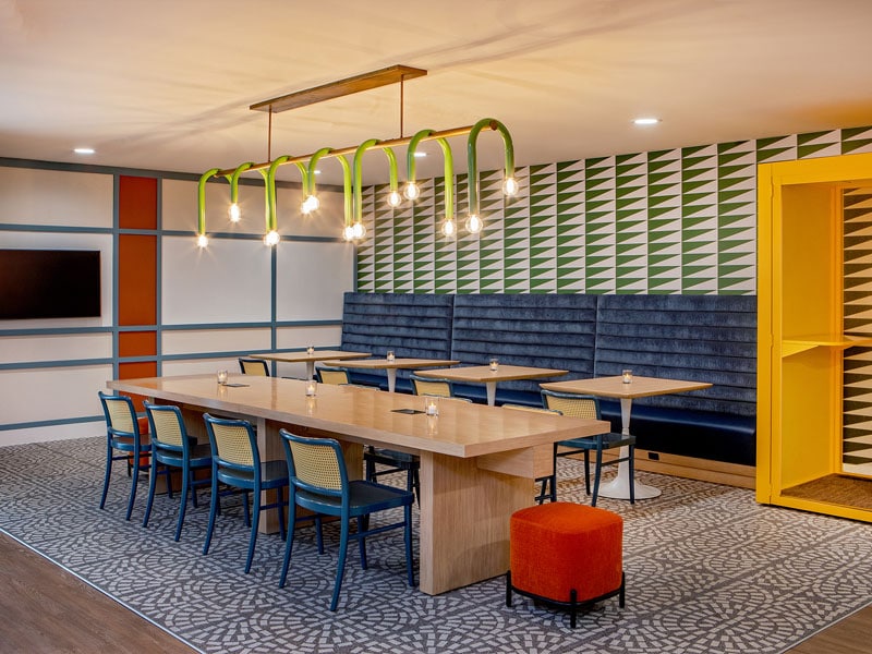 A spacious dining area features a long wooden table surrounded by colorful chairs. Bright lighting fixtures hang above, and vibrant wall patterns create a modern ambiance. A cozy booth with plush seating runs along one side, while a bright yellow door adds a pop of color.