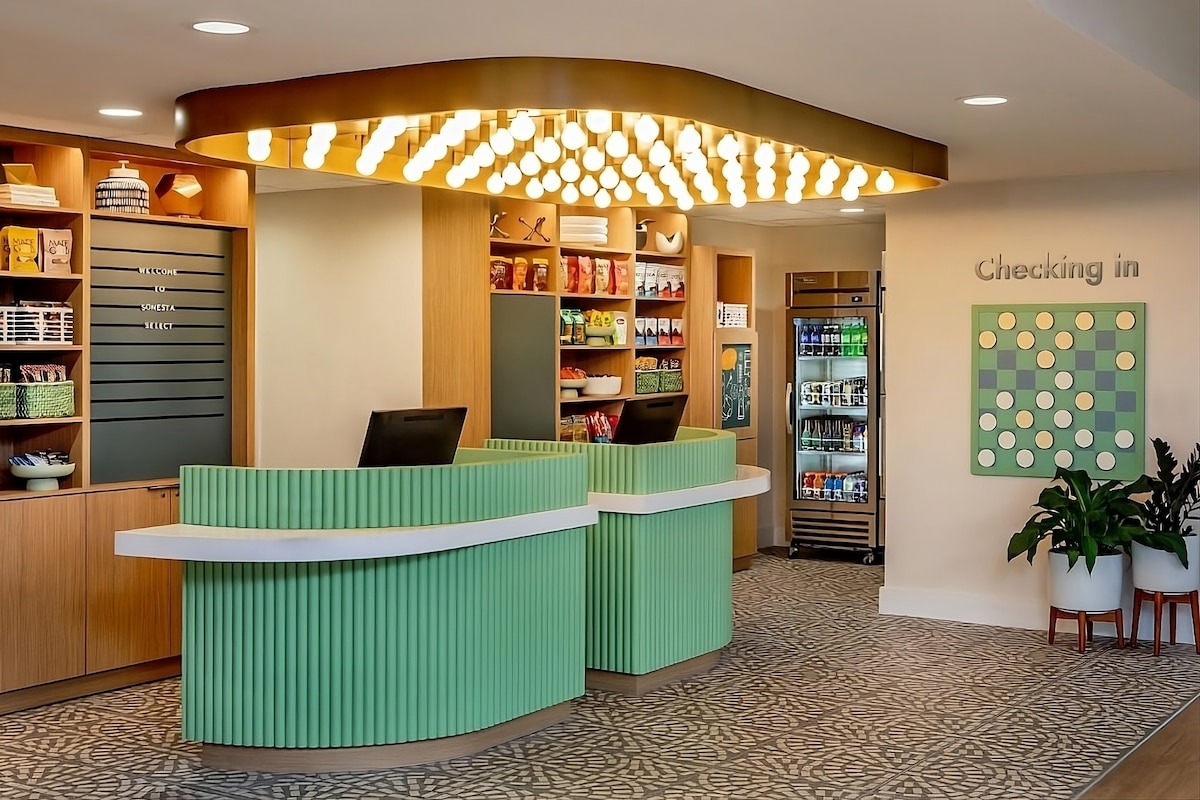 The hotel lobby features a modern design with green check-in desks, neatly arranged shelving, and a colorful display of refreshments. A striking light fixture adorned with multiple bulbs hangs overhead, creating an inviting ambiance. The space is complemented by decorative plants and a patterned floor.