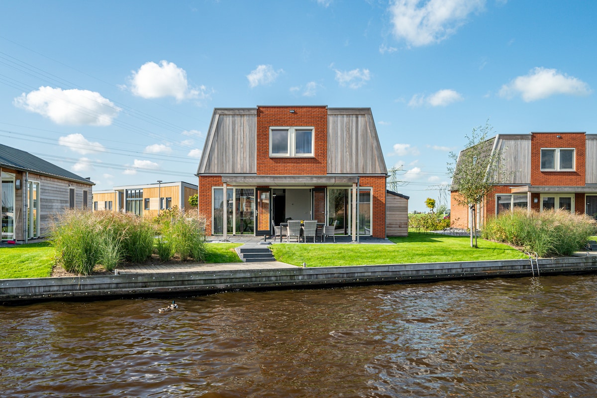 A modern two-story home sits by the water, featuring large windows and a wooden façade. A patio with outdoor furniture faces the lawn, surrounded by lush greenery. The private jetty extends into the water, providing easy access for water activities.