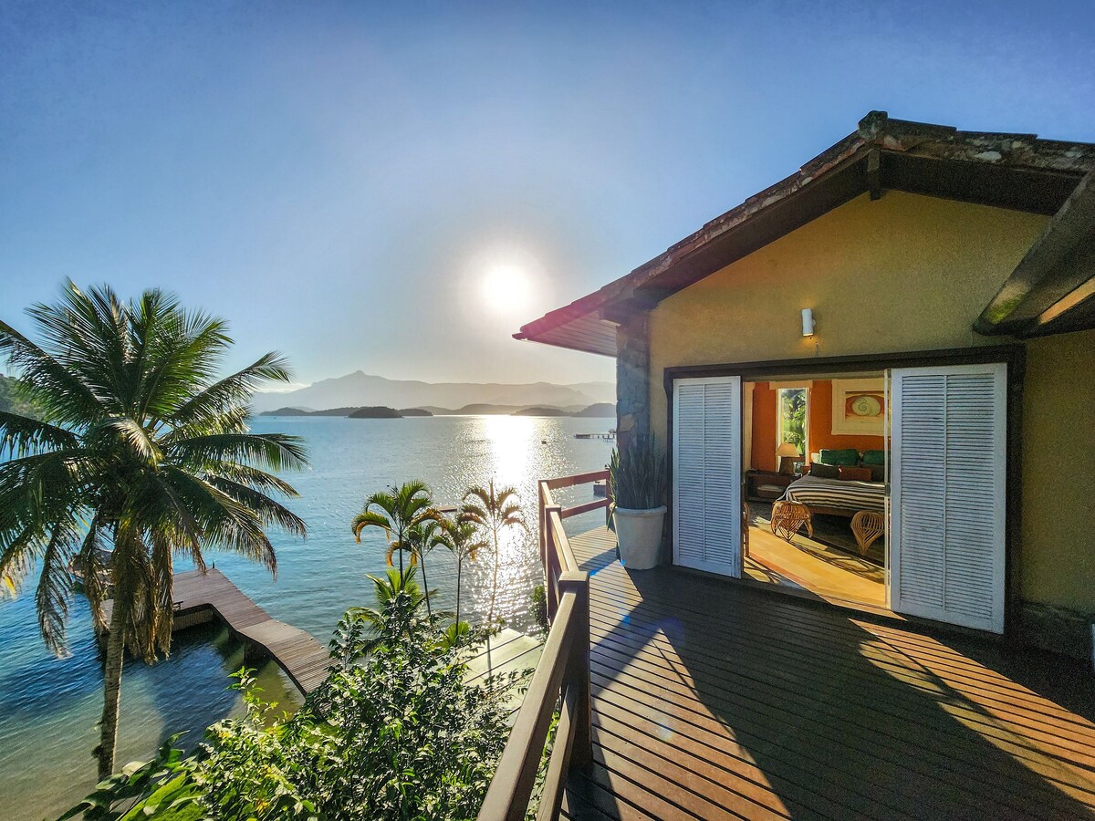 A sunlit view is captured from a deck overlooking a tranquil bay, with palm trees framing the scene. The water reflects the sunlight, and the landscape includes distant mountains under a clear sky. The open door reveals a glimpse of a cozy interior.