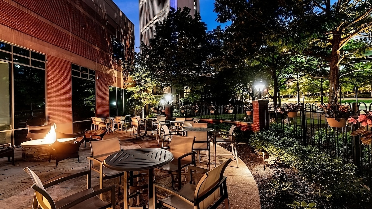 An outdoor space is featured, with several tables and chairs arranged on a stone patio surrounded by greenery. Soft lighting enhances the atmosphere, illuminating the area while trees and plants provide a natural backdrop.