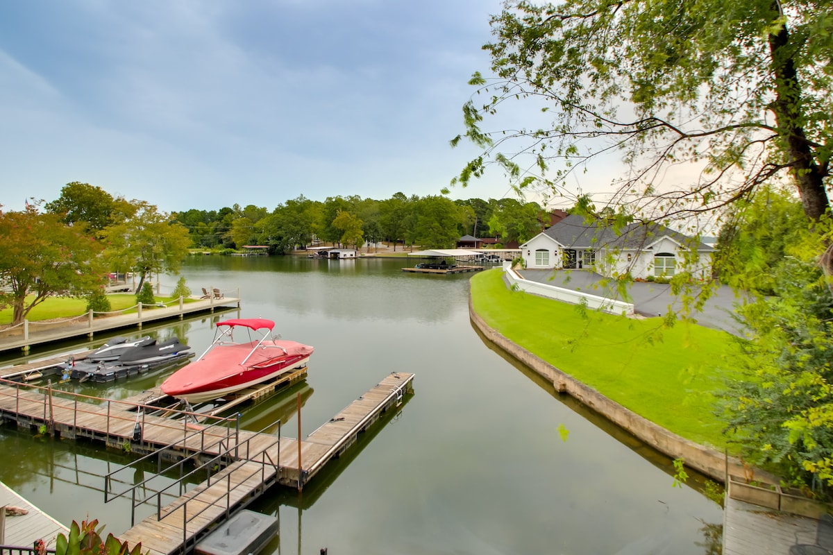 A serene view of the lake showcasing private boat docks. The calm waters reflect surrounding greenery and trees. In the foreground, a red boat is moored beside wooden docks, while the spacious property lies in the background against a soft, overcast sky.
