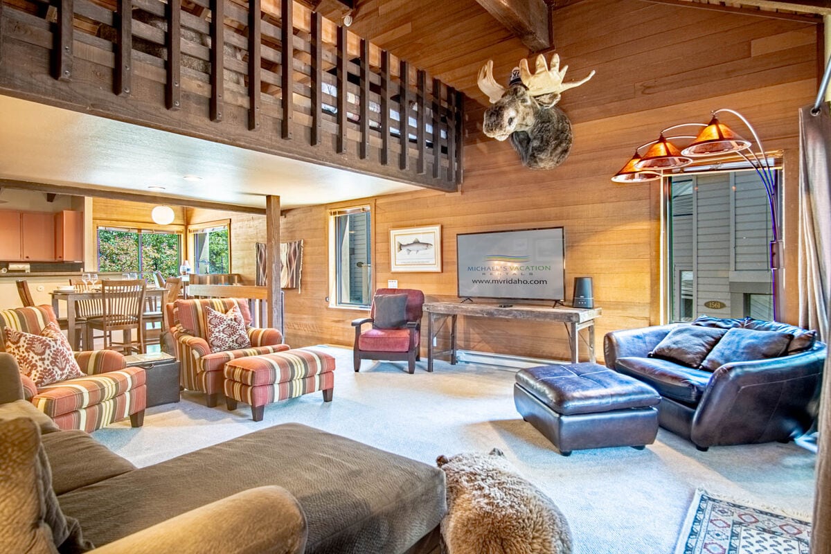 An inviting great room is captured with wood-paneled walls and vaulted ceilings. Cozy seating includes a striped armchair and a dark leather couch. A flat-screen TV is mounted on the wall, while natural light filters through large windows, enhancing the relaxed ambiance.