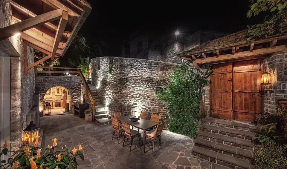 A charming outdoor space is revealed, featuring a stone courtyard illuminated by soft lighting. A wooden table with several chairs is positioned beneath a rustic wooden structure, while a decorative lantern adds ambiance. The surrounding stone walls showcase the villa's historic character.
