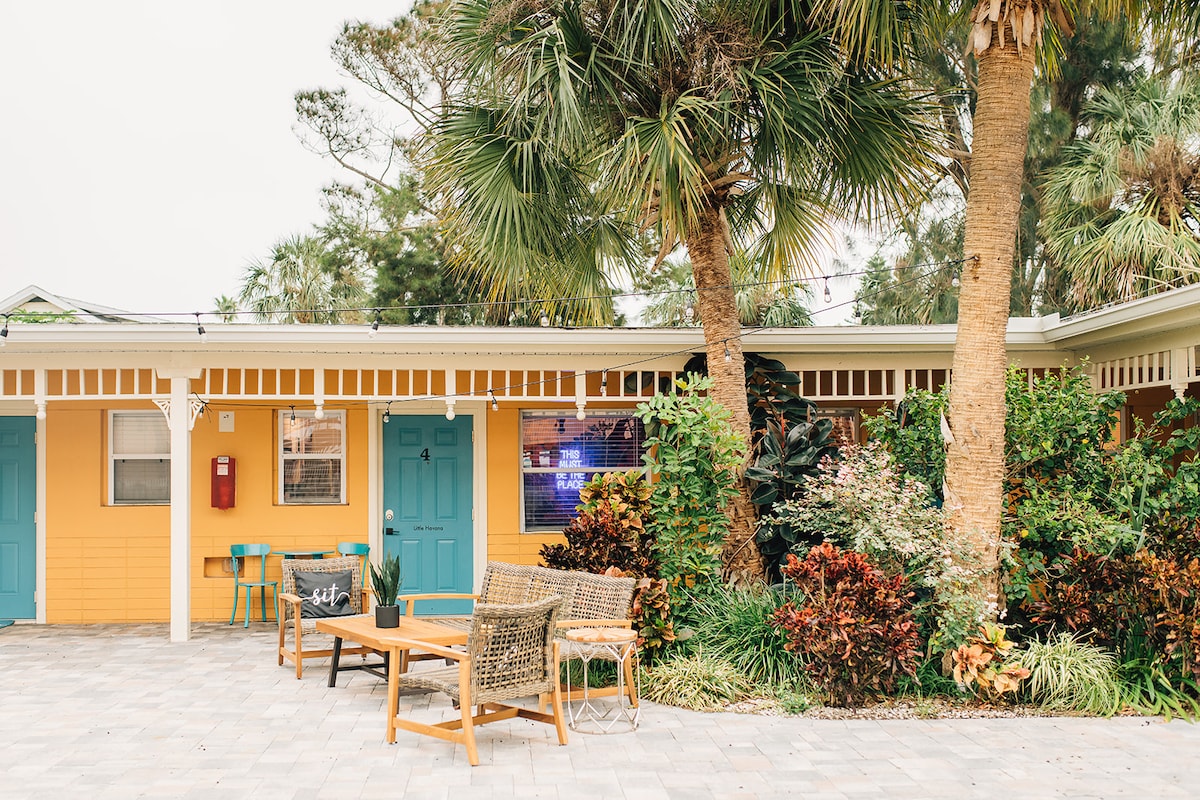 An outdoor seating area is surrounded by lush greenery and palm trees. A wooden table and chairs are set on a stone-paved patio, featuring vibrant foliage and colorful landscaping that creates a welcoming environment.