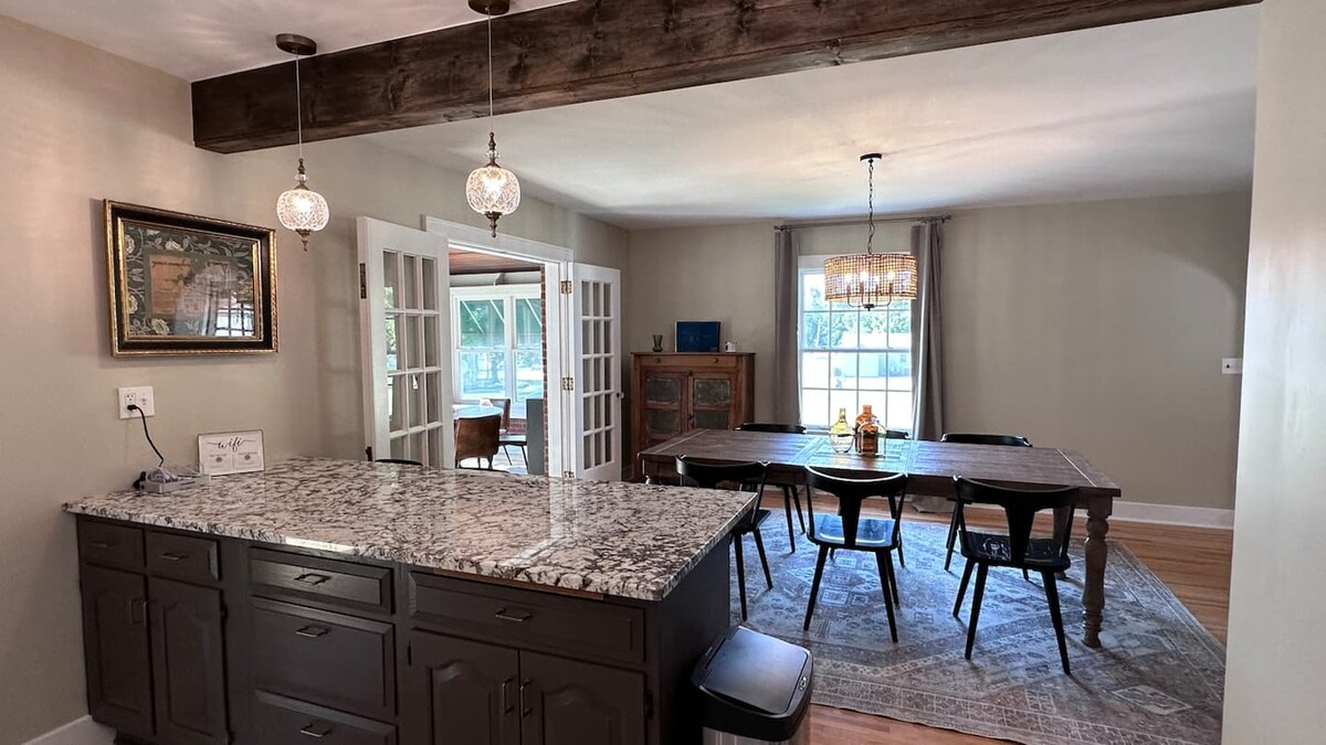 A spacious dining area is visible, featuring a long wooden table surrounded by black chairs. Natural light floods the room through large windows, complementing the warm tones of the walls and furnishings. A granite countertop is seen in the foreground, adding a modern touch to the space.