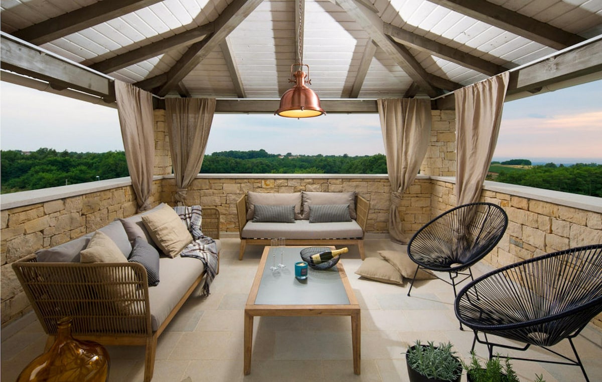 A spacious terrace is adorned with natural stone walls and exposed wooden beams. Comfortable seating areas include a sofa and two stylish chairs. Soft curtains frame the open space, providing a gentle breeze and an unobstructed view of the surrounding greenery.
