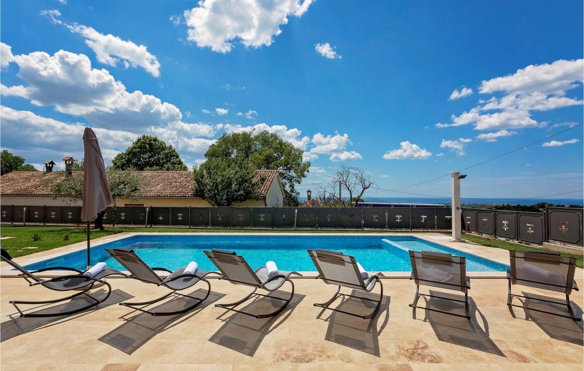 A spacious outdoor area features a swimming pool surrounded by sun loungers, each equipped with rolled towels. A backdrop of greenery and a distant view of the sea is visible under a clear blue sky with scattered clouds.