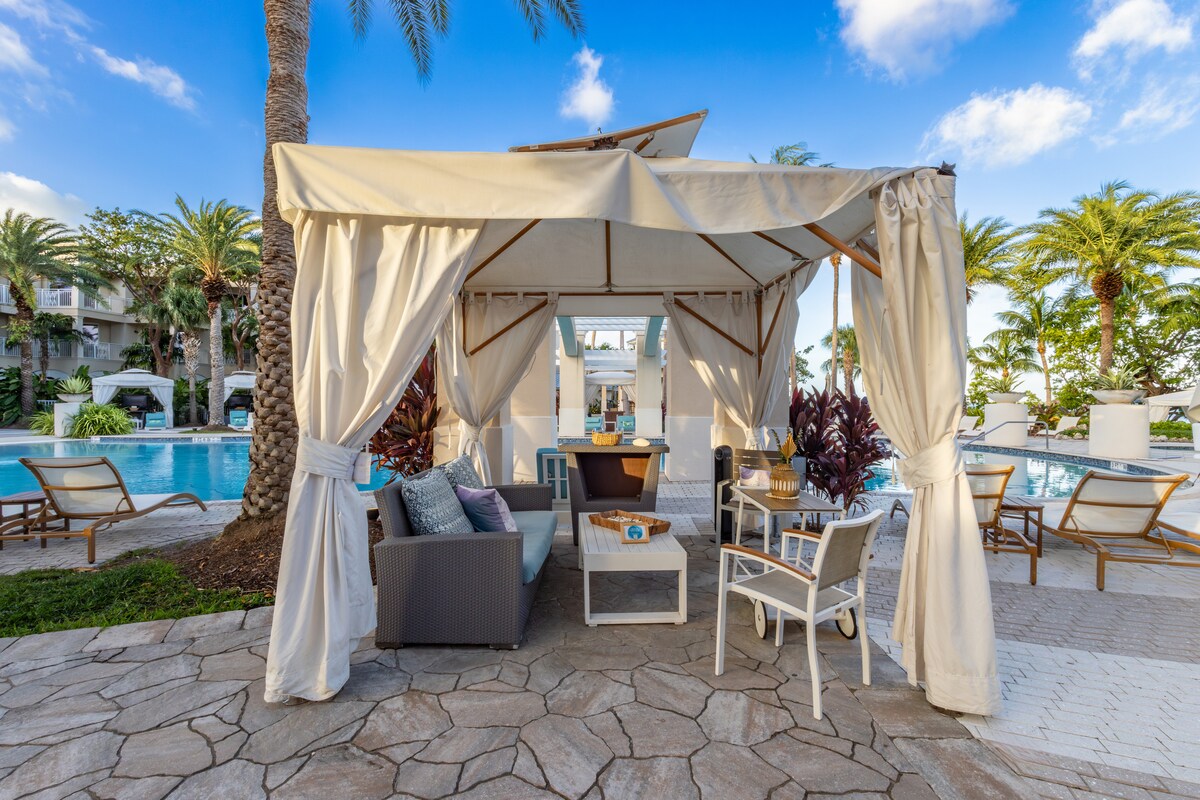 A shaded cabana near the pool features comfortable seating, including a cozy sofa and chairs. Lush tropical plants surround the area, with palm trees in the background. Soft lighting is visible, creating a relaxed atmosphere for gatherings or quiet moments by the water.