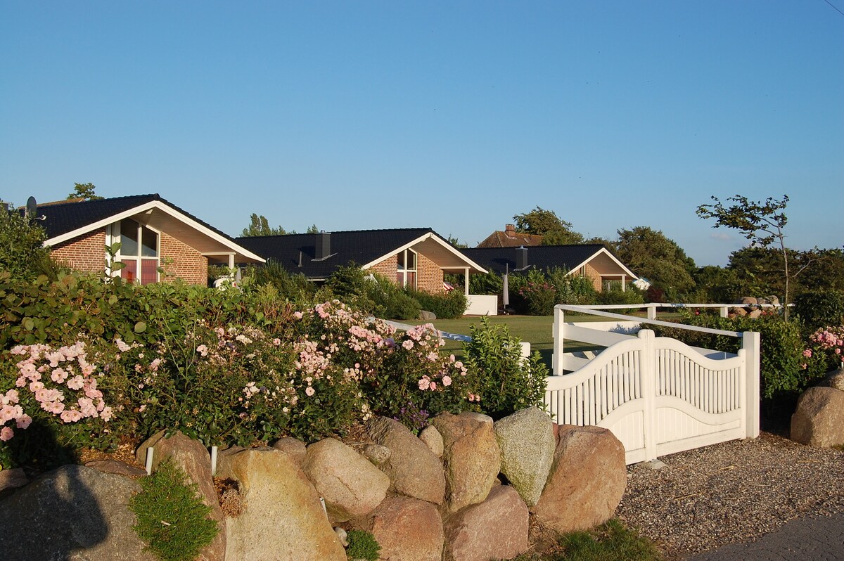 A serene view of several vacation houses is framed by blooming rose bushes and large stone borders. The houses, featuring sloped roofs and large windows, are set against a clear blue sky, creating a tranquil and inviting appearance.