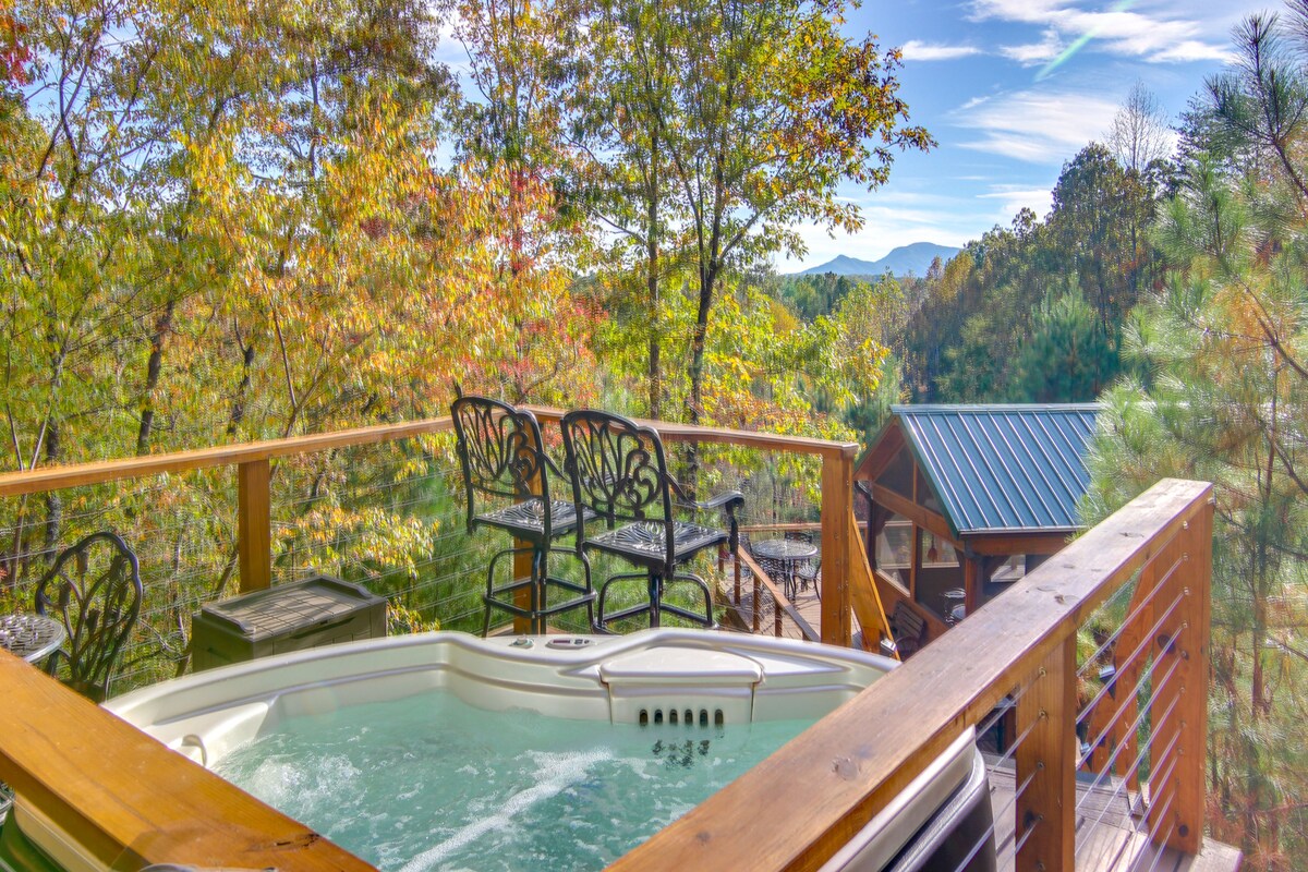 A private hot tub is positioned on a wooden deck overlooking scenic mountain views. Black metal bar stools are placed nearby, surrounded by lush green trees that add to the tranquil setting. The clear blue sky complements the natural beauty of the landscape.