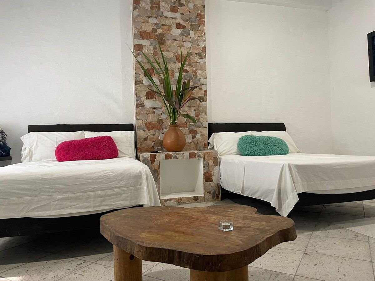 The sleeping area features two spacious beds with white linens, accented by colorful pillows in turquoise and pink. A wooden coffee table occupies the center, while a stone feature wall showcases a plant and adds texture to the serene atmosphere.