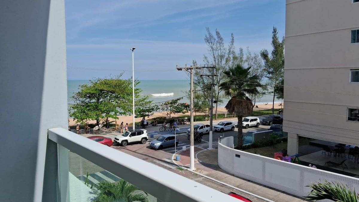 The image captures a balcony view overlooking a beach. The calm ocean waves can be seen in the distance, framed by tall trees lining the coastline. A few parked vehicles are visible on the street below, contributing to the scene's lively atmosphere.