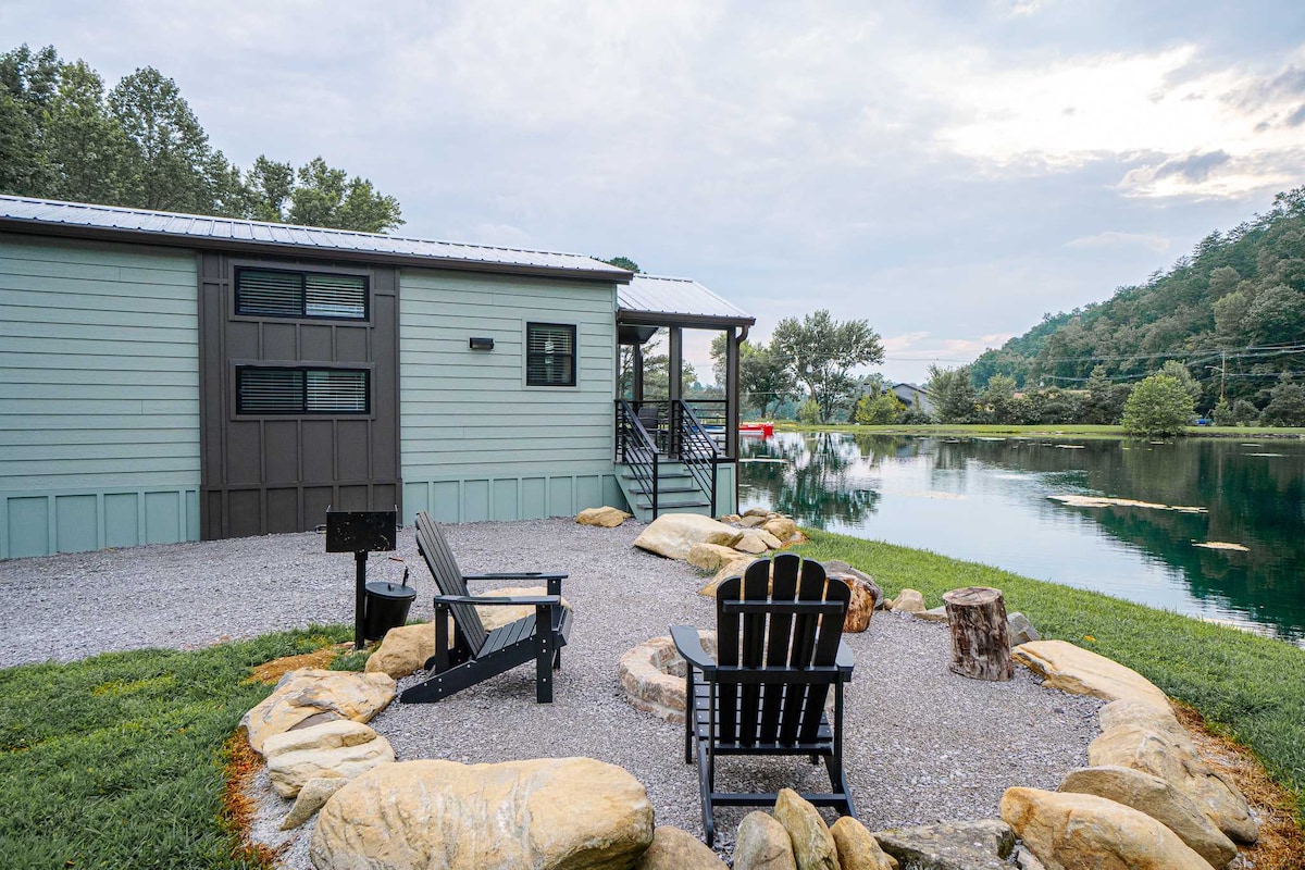 An outdoor sitting area is featured near a serene lakeside, with several adirondack chairs arranged around a stone fire pit. A gravel path leads to the entrance of the tiny home, framed by lush green grass and a gentle slope leading to the water.