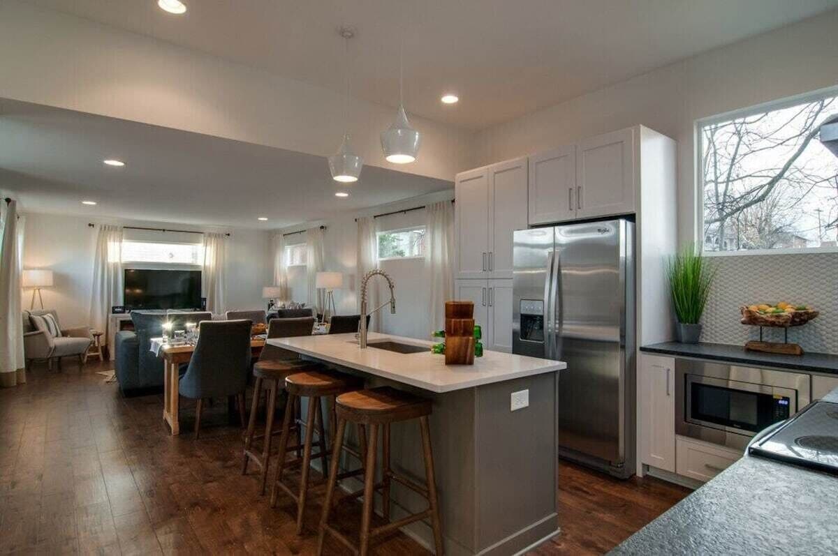 The open-concept kitchen features modern stainless steel appliances, a large island with seating for four, and under-cabinet lighting. Natural light fills the space through the nearby windows, creating a warm and inviting atmosphere. The living area is visible in the background, enhancing the spacious design.