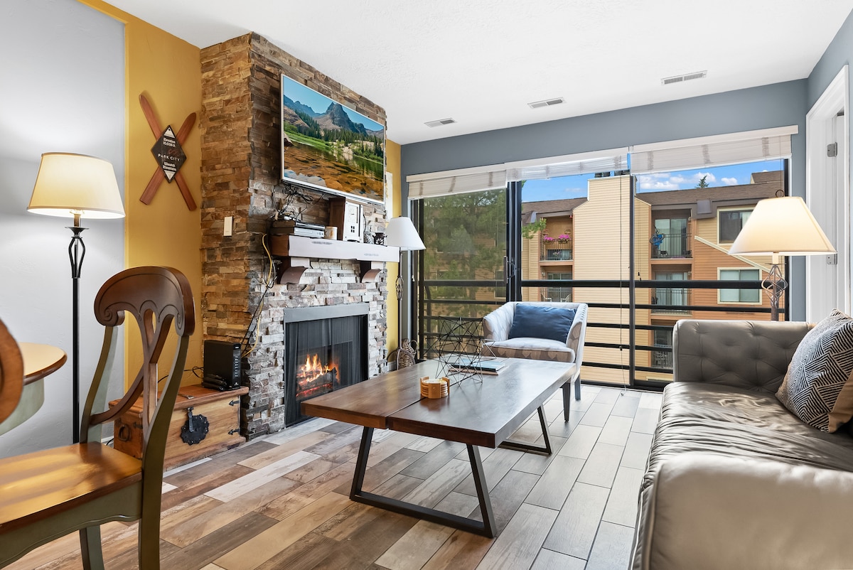 A welcoming living area features a stone fireplace at its center, flanked by a plush sofa and a wooden coffee table. Natural light streams in through the large sliding doors, offering views of the outdoors. A dining table with chairs is located beside the fireplace.