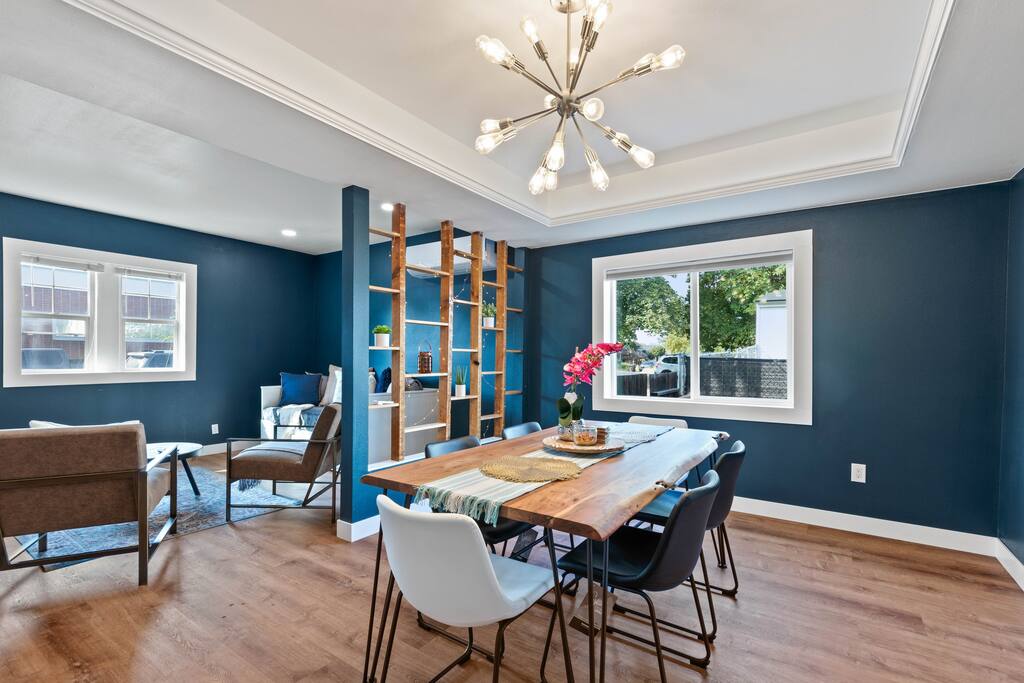 An open dining area features a wooden table surrounded by six chairs. The wall is painted deep blue, complemented by large windows that invite natural light. A modern lighting fixture hangs from the ceiling, and a decorative bookshelf with plants adds a homey touch.