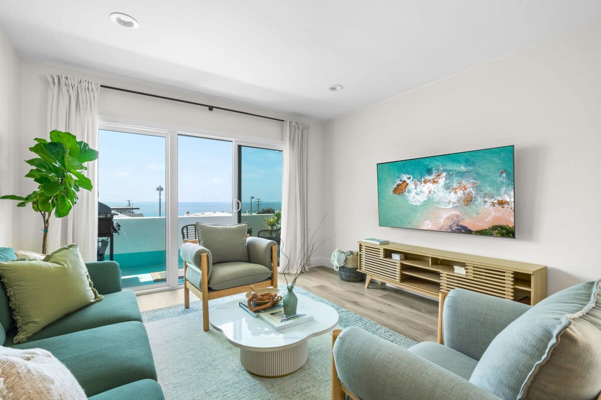 A well-lit living room features a comfortable sofa and armchairs, inviting relaxation. A coffee table sits in the center, and a large smart TV displays a vibrant ocean image. Natural light streams through sliding glass doors, revealing ocean views beyond.