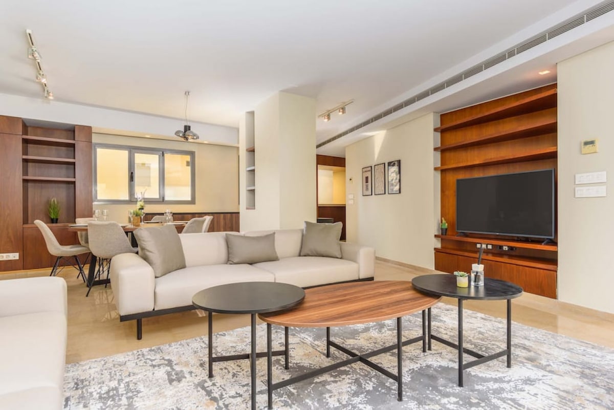 A spacious living area features a light-colored sofa, paired with contrasting dark coffee tables. The area is designed with minimal decor, allowing for natural light to brighten the space. Shelves are positioned on the walls, and a television is mounted for entertainment.