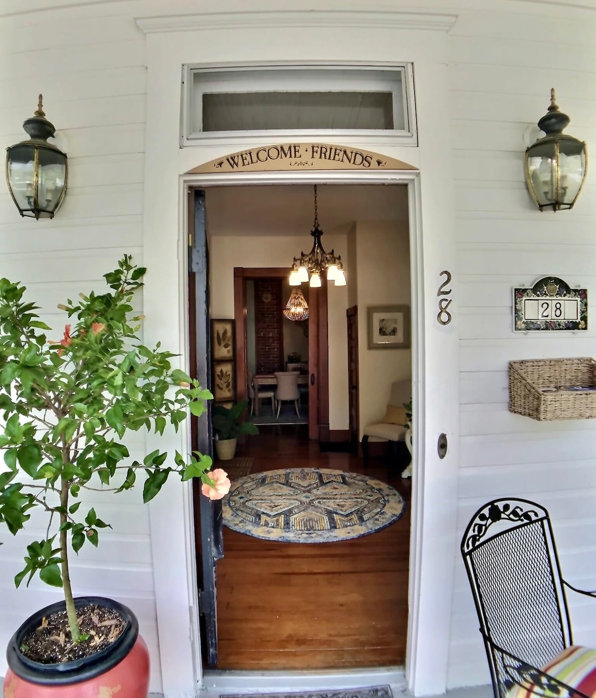 An inviting entrance features a charming door adorned with a welcome sign. Two elegant wall sconces flank the doorway, illuminating the path. A round patterned rug lies on the wooden floor, leading into a cozy common area visible through the doorway.