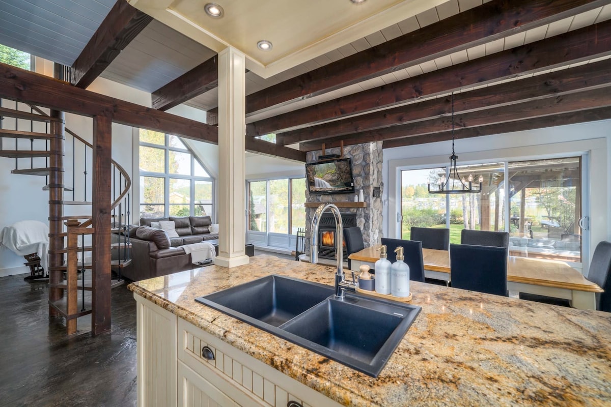A spacious kitchen is showcased with a large granite island and modern sink. Exposed wooden beams add character, while natural light floods through expansive windows, revealing a cozy seating area and a dining space in the background, creating an inviting atmosphere.