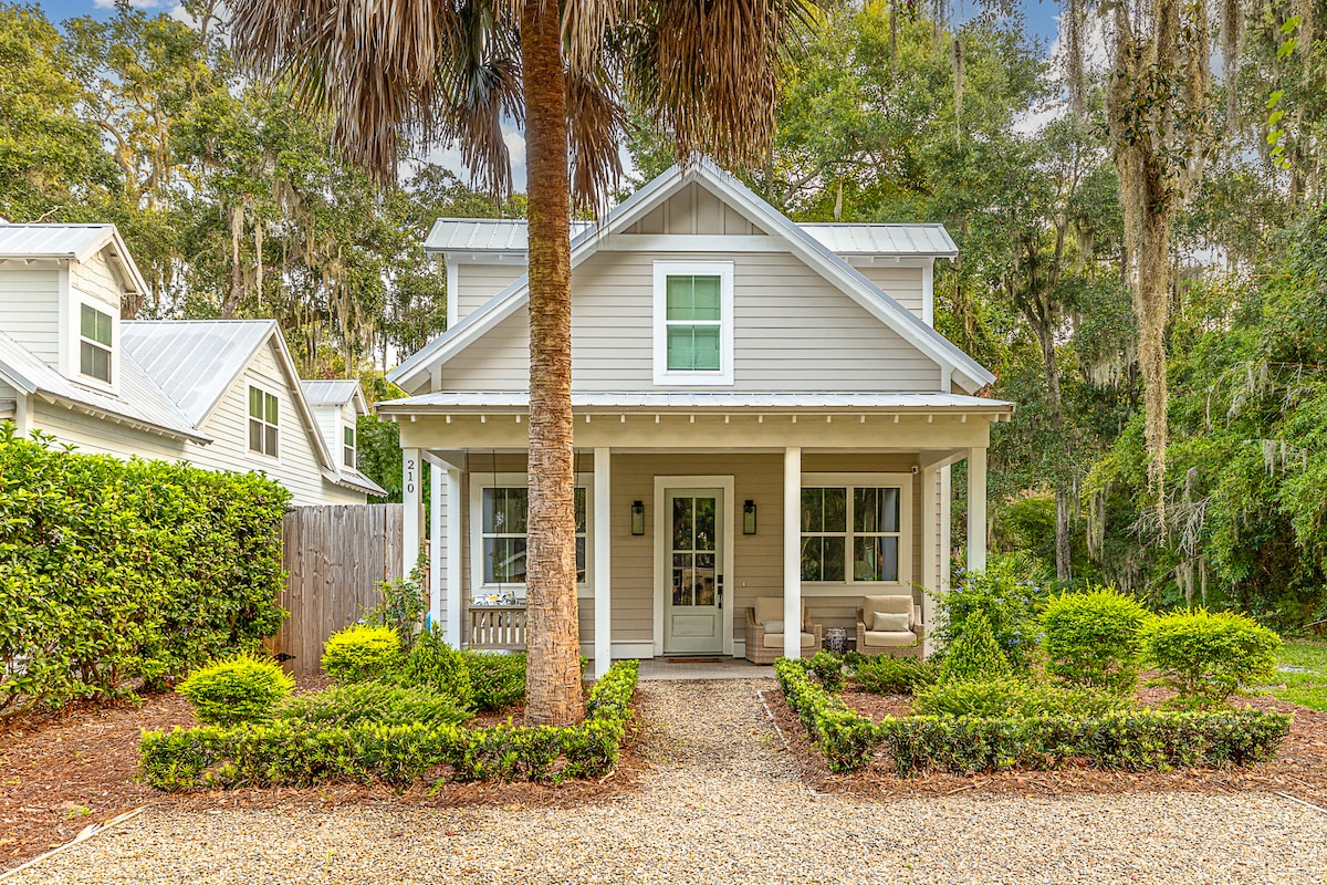 The exterior of a charming home is showcased, featuring a light gray facade and a covered front porch with two seating chairs. Lush greenery frames the property, including shrubs and palm trees. A pebbled driveway leads to the entrance, creating a welcoming approach.