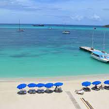 A serene beach view captures light sand lined with blue umbrellas over lounge chairs. Calm turquoise waters stretch towards the horizon, dotted with boats. A wooden dock juts out into the water, enhancing the tranquil coastal atmosphere.