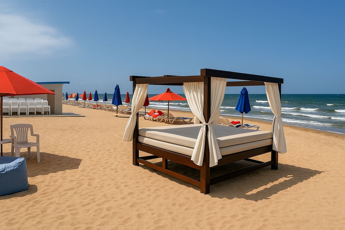 A shaded beach cabana with draped fabric stands on golden sand, offering a cozy spot for relaxation. In the background, colorful umbrellas and loungers provide ample seating, while gentle ocean waves create a serene coastal atmosphere under a clear blue sky.