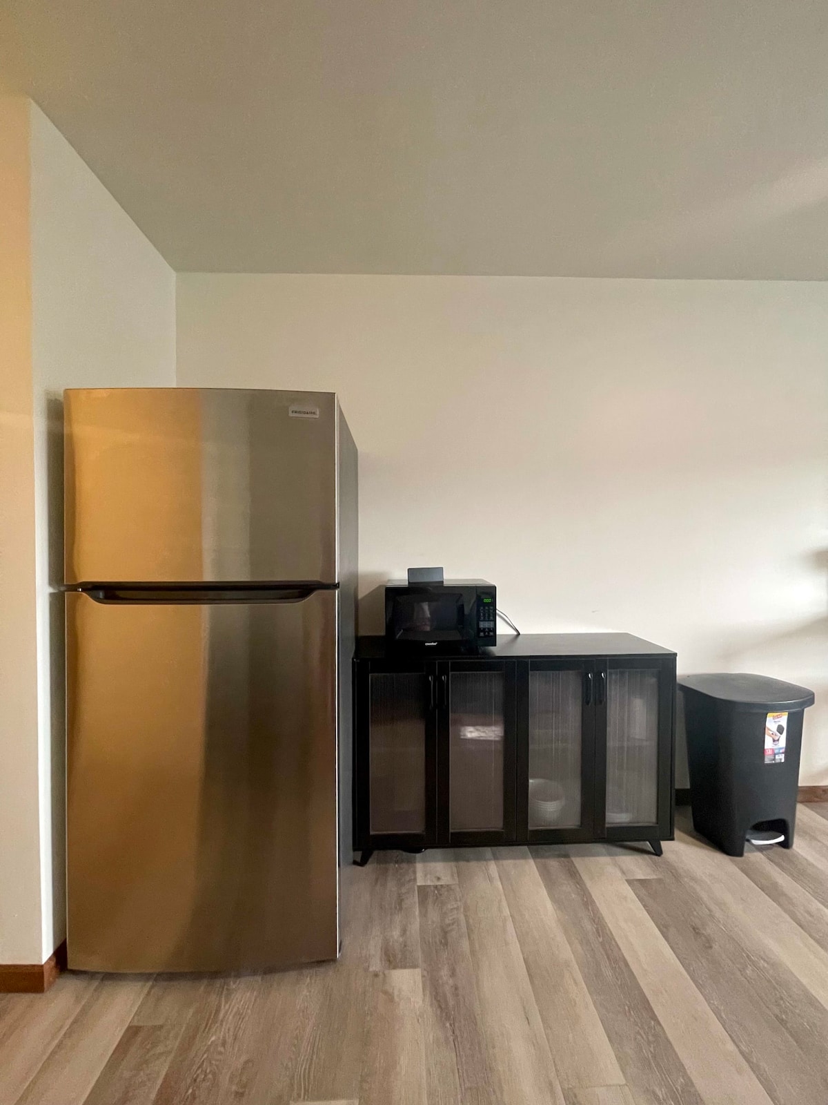 A modern kitchenette area is shown, featuring a stainless steel refrigerator beside a microwave and a cabinet with glass panes. A waste bin is positioned nearby, all situated on brand new, light-colored LVP flooring. Natural light highlights the sleek design and updated appearance.
