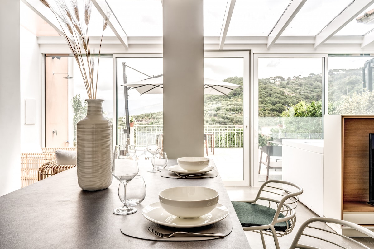 A modern dining area is equipped with a minimalist table set for four, complete with elegant dinnerware and glasses. Large glass doors invite natural light and highlight views of the surrounding hillside and terrace, seamlessly connecting indoor and outdoor spaces.