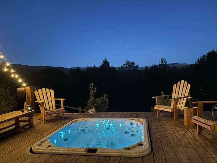 A hot tub is situated on a wooden deck, surrounded by Adirondack chairs. Soft lighting illuminates the area, contributing to a serene ambiance. The distant hills are visible against the twilight sky, enhancing the peaceful outdoor setting.