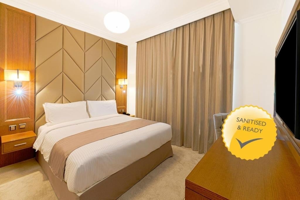 A spacious bedroom features a plush double bed with neutral bedding. Textured walls create visual interest, while soft curtains frame the large window. A flat-screen TV and wooden furniture complement the contemporary design, all illuminated by warm lighting.