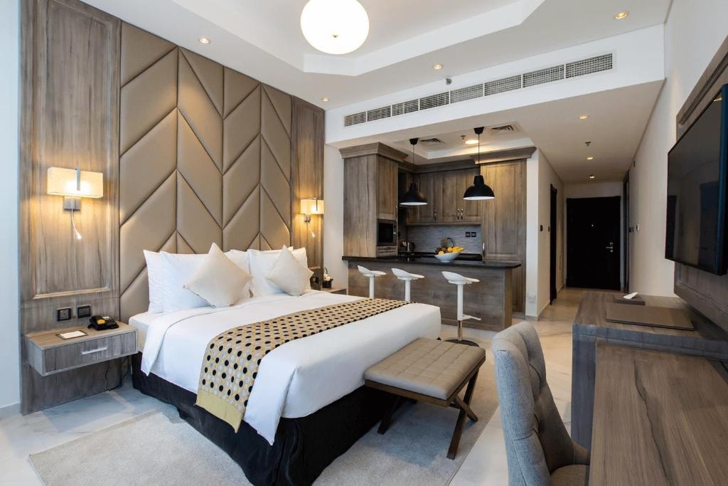 The spacious bedroom showcases a large bed with stylish bedding, complemented by subtle wall paneling. A kitchenette with bar seating and modern appliances is visible. Soft lighting accents the room's decor, while a seating area with a desk provides additional functionality.