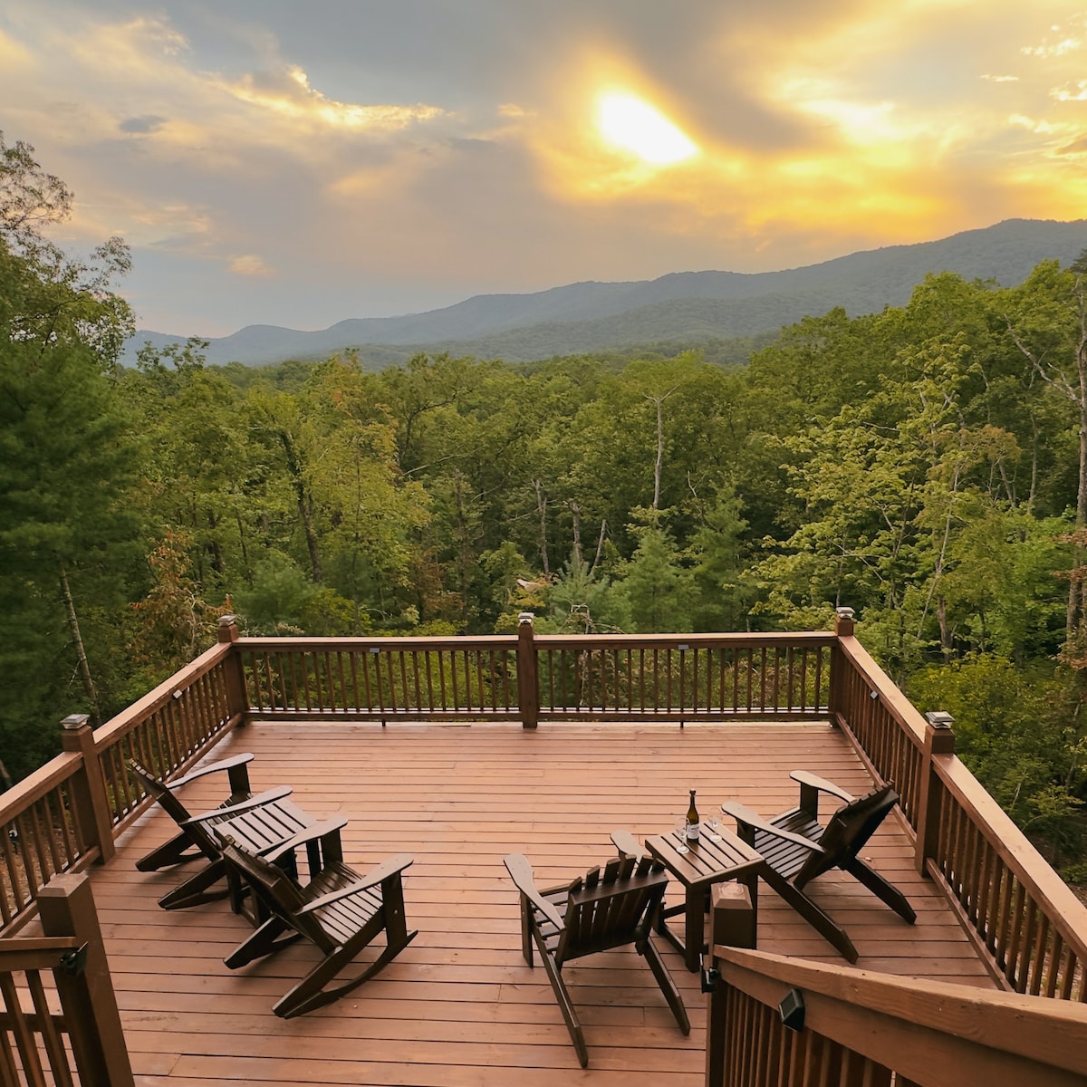 An expansive wooden deck features several Adirondack chairs, offering a perfect spot for relaxation. The surrounding trees and distant mountains are illuminated by a soft sunset, creating a serene outdoor setting.
