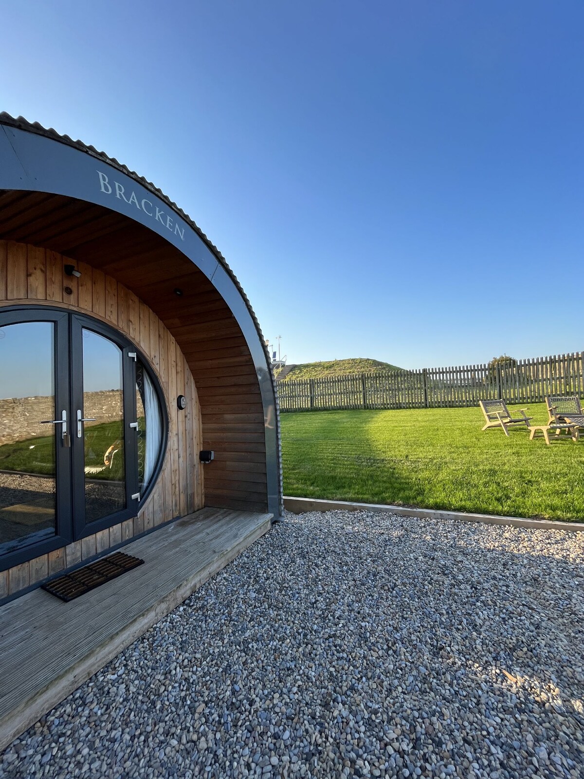 The exterior of the Bracken pod is shown, featuring a curved wooden structure with double glass doors. A gravel path leads to the entrance, which is framed by a grassy area. Garden furniture is visible in the enclosed space, under a clear blue sky.